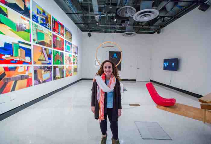 A short-statured woman with long brown curly hair and brown eyes stands in the middle of a white cube gallery space, smiling at the camera. She is wearing red lipstick to match the red and white scarf wrapped around her neck. On every wall of the gallery space hangs contemporary art that includes brightly-colored photographs of abstract shapes, a round wooden ring hanging from the ceiling, a black and white video installation of a sign language interpreter and a low-lying red lounge chair on a mat. The ceiling is exposed so all the beams, wiring and fan ducts are visible.