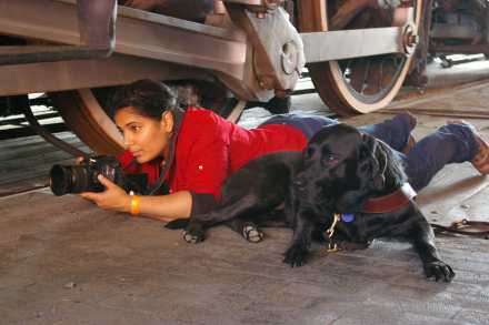 An Arab-American woman with dark hair in jeans and a red shirt on the floor holding a camera about to take a photo. Next to her lying on the ground is a black Labrador guide dog.