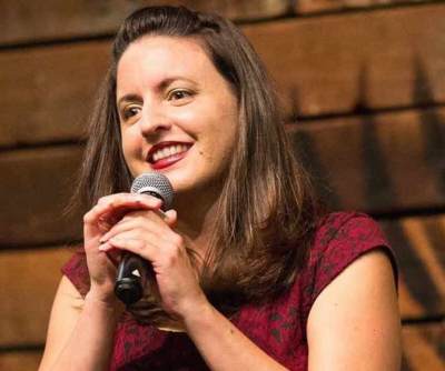 Nina G, a young white Italian American woman with long brown hair and a short-sleeved red dress smiling and holding a microphone.