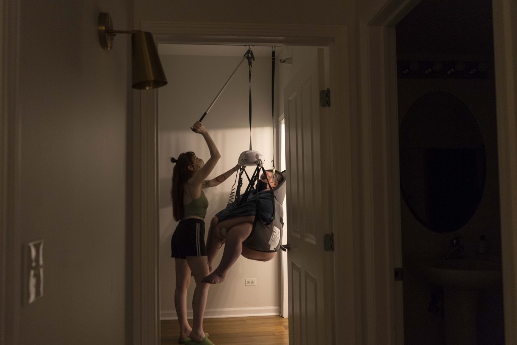 A landscape, side-profile photo of Marley moving Kennedy through the room using a ceiling hoyer lift. Facing each other in masks, Marley and Kennedy are framed by a doorway as Marley reaches up to operate the lift Kennedy is suspended in. Warm light pours in from the bathroom door behind them, though the rest of the surroundings are dim. Kennedy holds the remote operating the lift, her butt and legs hang down from the sling.