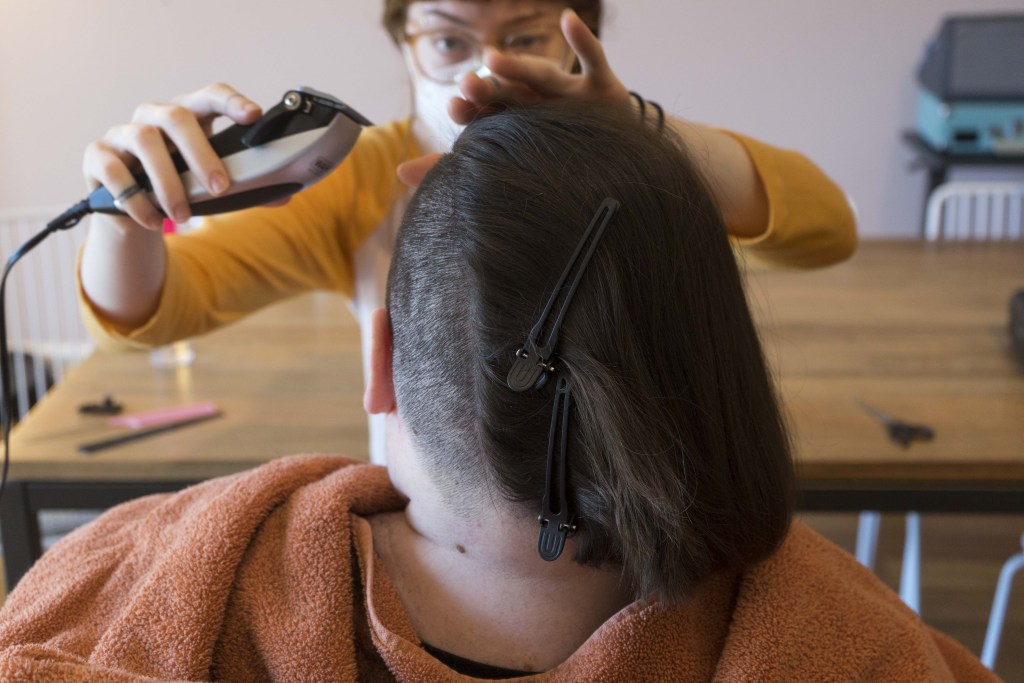 A close-up landscape photo of Marley giving Kennedy a haircut. Kennedy’s head is seen from behind, with long hair on the right side of her head sectioned off with clips. Marley faces the camera, peering over Kennedy’s head, re-shaving the left side of her hair with clippers. There is an orange towel around Kennedy’s shoulders and tools for cutting hair are blurred on the wood table behind them.