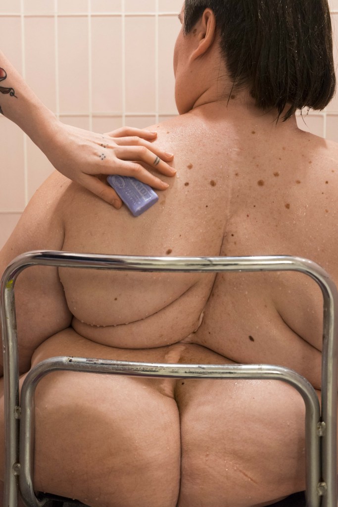 A portrait photo of Kennedy in the shower as Marley washes her back. Kennedy is facing towards the pink tiled walls of the shower with the back of her head, back, and butt filling the frame. She has a long scar down the center of her back and multiple back fat rolls. A metal chair back frames her body. Marley’s hand reaches in from the left with a purple bar of soap.
