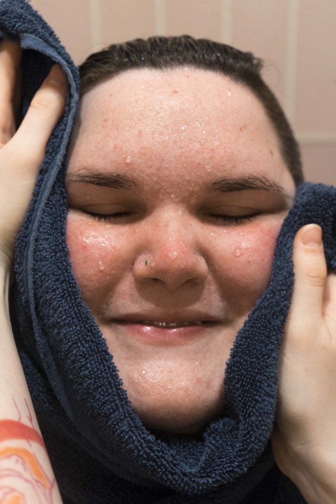 A portrait photo of Kennedy’s face filling the frame as Marley’s hands surround it with a blue towel. Kennedy’s eyes are closed, mid-smile, her face dotted with water droplets. A tattoo is visible on Marley’s left arm.