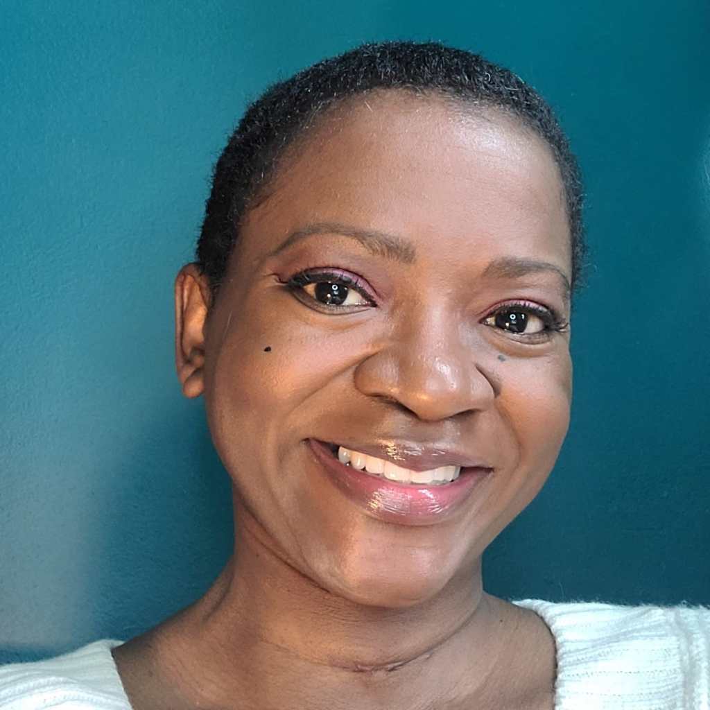 Photo of a Black female with dark brown complexion, close cropped black hair, wearing a wide smile, and white sweater, in front of a teal colored wall.