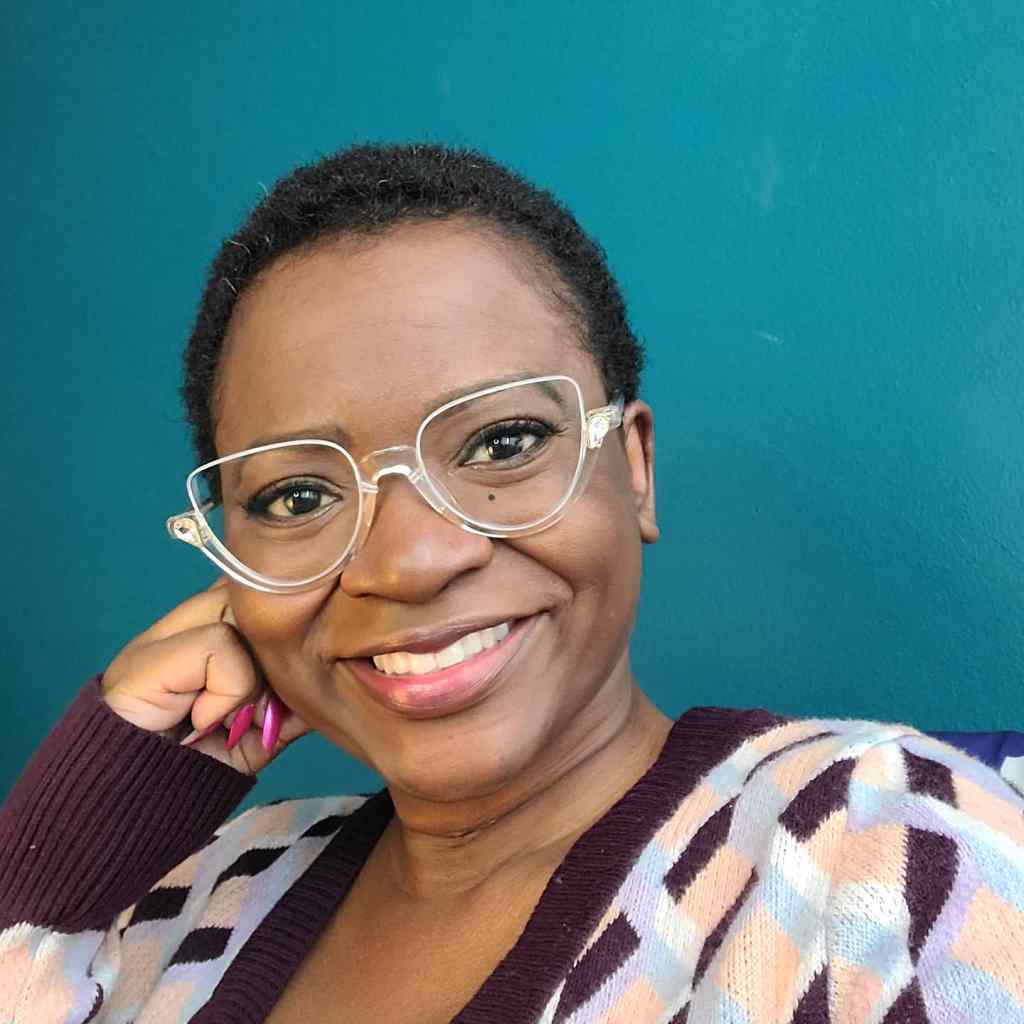 Photo of a Black female with dark brown complexion, close cropped black hair, wearing a wide smile, clear plastic glass frames and sweater with a geometric pattern in eggplant, sky blue, white and light peach colors.