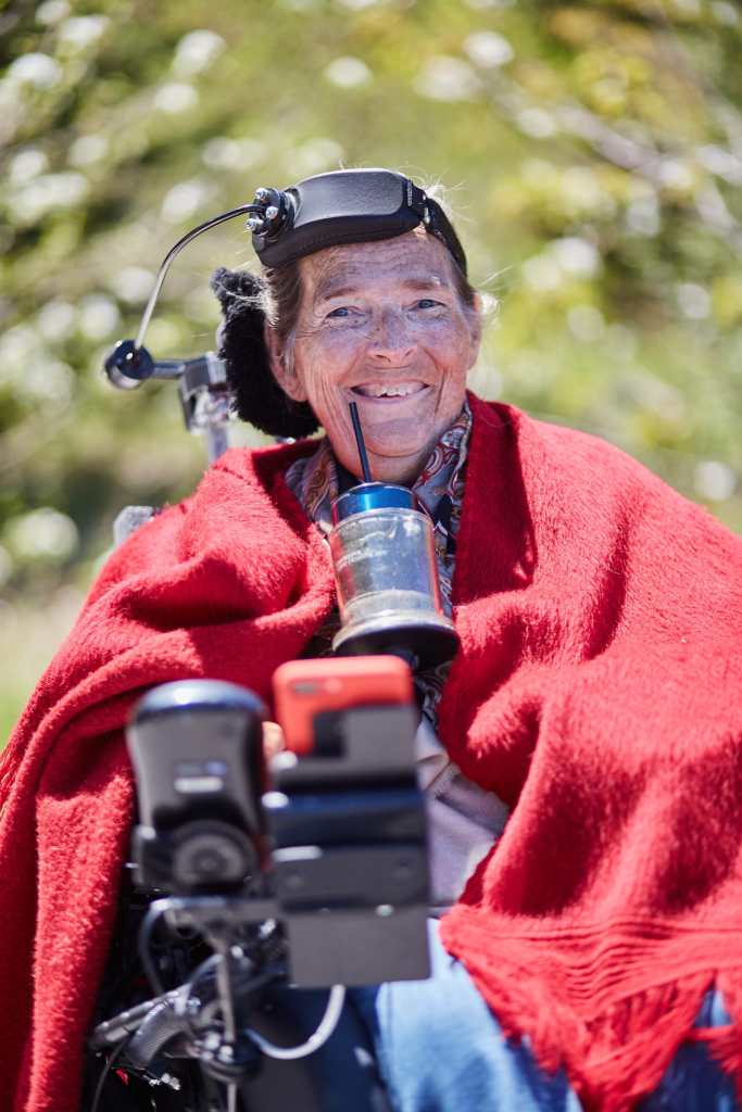 Catherine, a white woman, smiles widely like the joyous elder crip lesbian she is. She is seated in her power chair wearing a red shawl with a blur of lush greenery behind her.