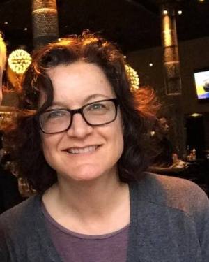 Photo of Claudia Center, a white woman with wavy shoulder-length brown hair. She is wearing glasses and a heather gray cardigan with a purple t-shirt underneath. She is smiling at the camera.