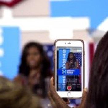 Photo of a person holding a smartphone taking a photo of First Lady Michelle Obama at a rally.