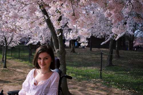 A young white woman with chin-length brown hair smiles at the camera. She wears a white shirt and sits in a black motorized wheelchair. Behind her are several blooming cherry blossom trees.