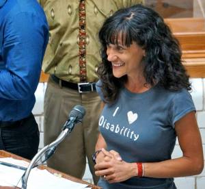 Image description: A white woman with long, curly hair smiles broadly, with raised eyebrows, and speaks into a microphone on a podium. She clasps her hands together tightly and looks out at her audience. Her t-shirt is decorated with 