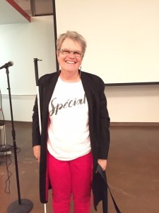 A white woman with short hair is standing with her white cane by her side. She is wearing a long black jacket, red pants and a white shirt that says, 'Special.' She is standing behind a classroom with a white board in the back.