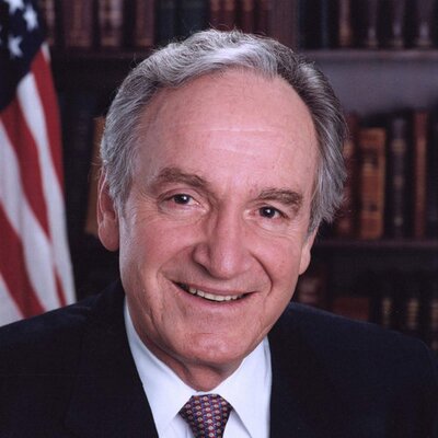 Photo of Senator Tom Harkin. He has short white hair and wearing a dark suit with a white shirt. An American flag and a bookshelf full of books is in the background