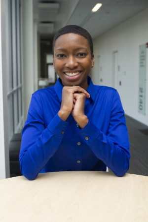 Image of a young genderfluid black person of African descent (TL) wearing a bright blue collared shirt while seated at a desk in a hallway with their hands folded under their chin and elbows leaning against the tabletop. There are windows & doors in the background.