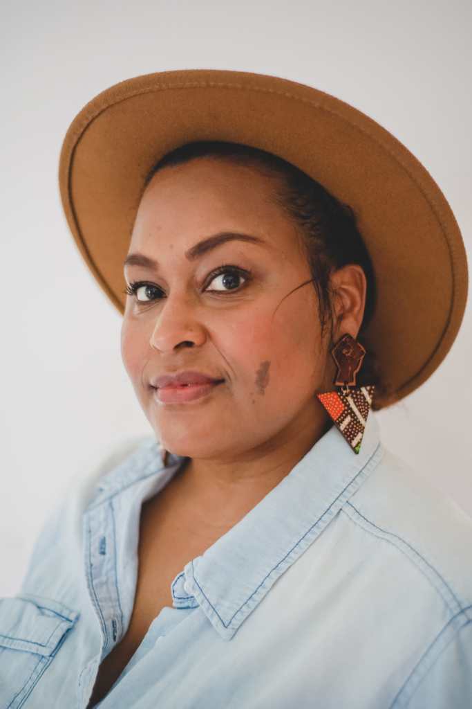 Photo of Jen White-Johnson, an Afro-Latina woman who is wearing a light blue denim shirt. She has a cinnamon complexion with a small brown birthmark on her left cheek. She is wearing earrings with the raised solidarity fist with an inverted triangle below. She is also wearing a wide-brimmed brown hat.