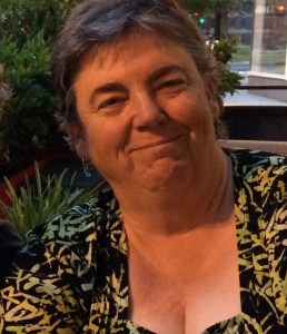 Photograph of Corbett Joan OToole, a light skinned female presenting person. Corbett has short greying hair, wears a black and yellow blouse and is smiling.