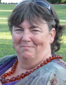 An older white woman with brown hair with bangs on her forehead. She has a pair of sunglasses on her head and is wearing a beaded necklace and a multicolored shirt. In the background is a grassy lawn.