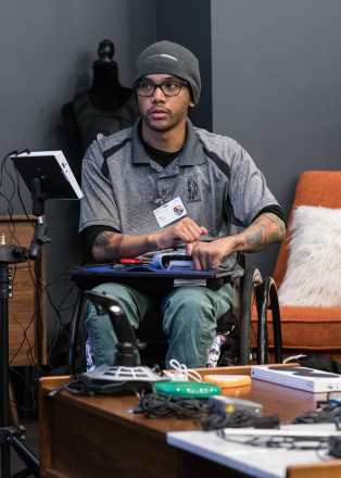 Photo courtesy of Microsoft. A person using an Xbox Adaptive Controller while gaming who appears to be a young man of color in a wheelchair wearing glasses, a gray short-sleeved shirt, green pants, and a gray knit cap.
