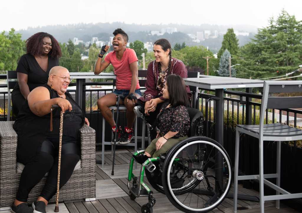 Five disabled people of color with canes, prosthetic legs, and a wheelchair sit on a rooftop deck, laughing and sharing stories. Greenery and city high-rises are visible in the background. Photo credit: Disabled and Here https://affecttheverb.com/disabledandhere/