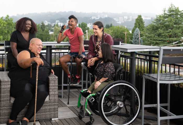 Five disabled people of color with canes, prosthetic legs, and a wheelchair sit on a rooftop deck, laughing and sharing stories. Greenery and city high-rises are visible in the background. Photo credit: Disabled and Here https://affecttheverb.com/disabledandhere/
