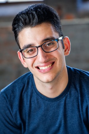 A headshot in which Ryan, a brunette with glasses, wears a navy blue t-shirt and smiles for the camera. 