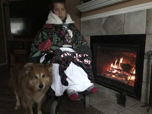 Day 2: Emily Wolinsky sits still next to her fireplace in a dark room. She’s covered in blankets of different colors and patterns. She has thick socks on her feet. Her dog, Byron, a fluffy sweet poof, sits next to her.