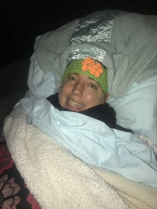 Emily Wolinsky lies in bed covered with head scarves, sweaters and blankets. She smiles wearily at the camera.