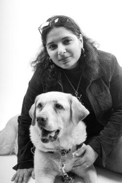 Image description: Black and white photo of an Arab-American woman with dark hair in jeans and casual wear, and yellow Labrador guide dog. Photo credit: Rachel Ellis
