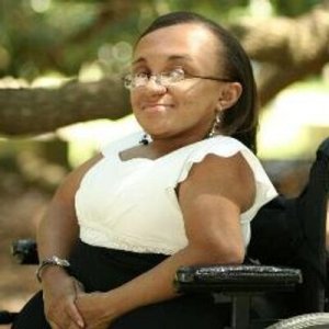 A young African American woman in a wheelchair. She's sitting outside with trees in the background. She's wearing glasses, a white sleeveless shirt and a black skirt.