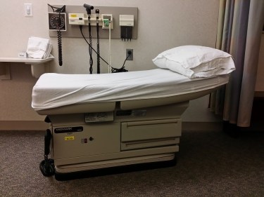 Picture of a typical exam table in a doctor's office that's covered in a white sheet with a white pillow. On the wall are various medical instruments such as a blood pressure cuff.