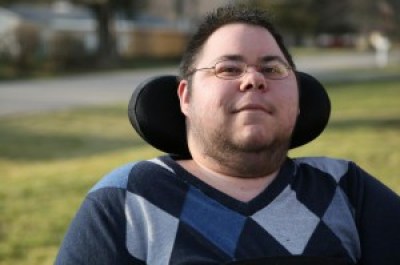 Photo of Dominick Evans, a transgender man with short dark hair and glasses. He has stubble around his chin and neck. He is in a wheelchair with a black headrest behind him. He is wearing an argyle-print sweater in different shades of blue.