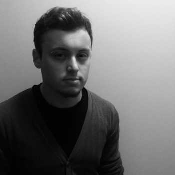 A black and white picture of Dominik, a white man with slightly curly brown hair, a dark pinch, and a piercing over the right eyebrow. He is wearing a cardigan over a black shirt.