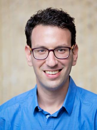 A headshot of a white man with dark hear and eyes smiling straight to the camera. He is wearing a blue colored shirt and square dark blue eyeglasses.