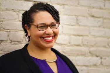 Medium shot of Dr. Sami Schalk. She is a light-skinned black woman with short curly hair and brown glasses. She is smiling to the camera in front of an off-white painted brick wall. She is wearing a black velvet jacket over a purple dress, red lipstick, and silver hoop earrings. Photo by Smoketree Photography of Madison, WI.