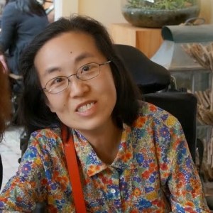 Image of an Asian American woman wearing glasses sitting in a wheelchair wearing a blouse with a flower print.