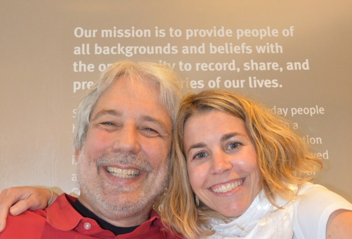 Photo of an older white man in a red shirt and using a wheelchair. He is next to a younger white woman with blonde hair. They are in front of a StoryCorps recording booth.