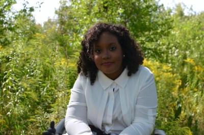 Image description: Black woman in a wheelchair sitting in front of trees looking directly into the camera with a smile.