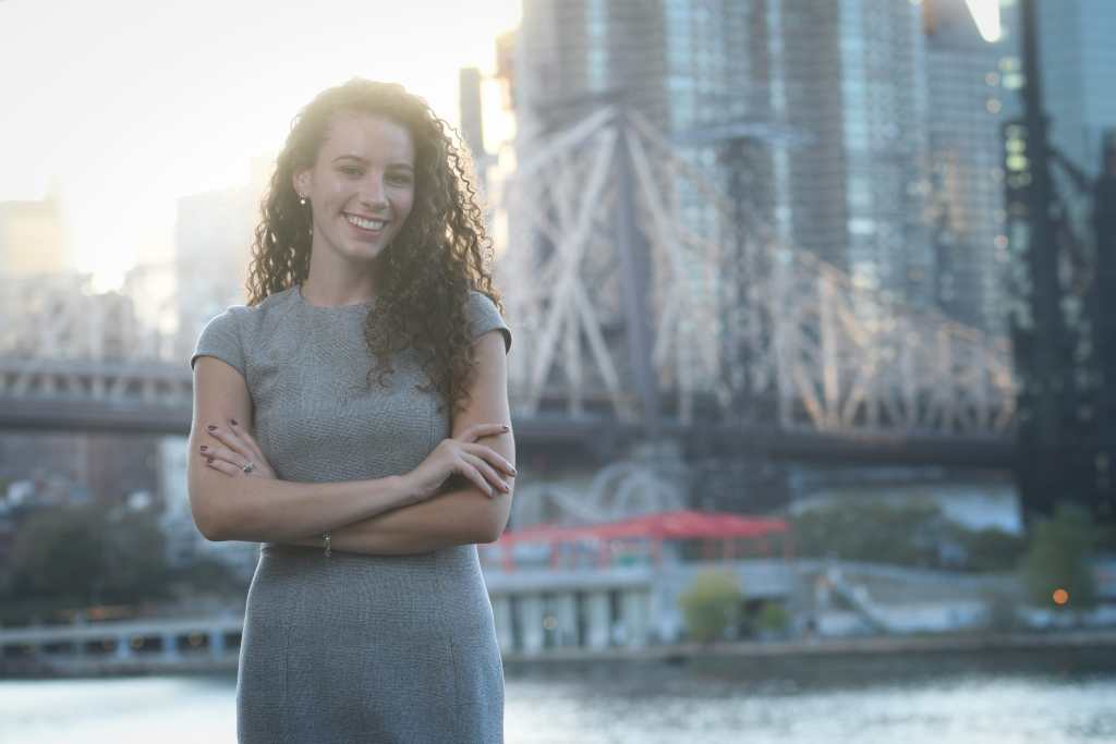 Rebecca Lamorte, candidate for New York City Council, is smiling and standing on Roosevelt Island in New York City in front of the Queensboro Bridge. Rebecca is a white woman with long, curly brown hair and is wearing a gray dress. 