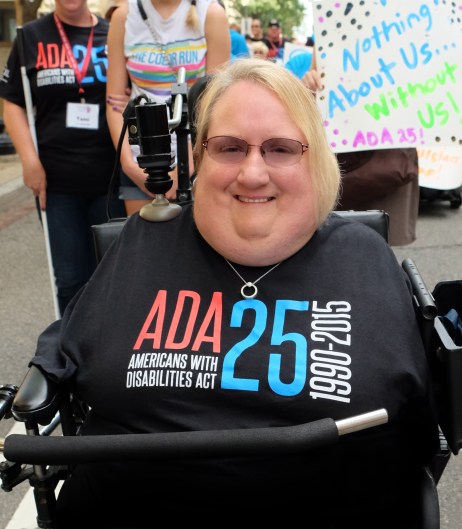 Marilee Adamski-Smith, a woman with short blond hair, is in a wheelchair with a bar across her torso. She is wearing a black t-shirt that says "ADA 25 Americans with Disabilities Act 1990-2015." She does not have any arms or legs and has a joystick resting on her right shoulder for her wheelchair.