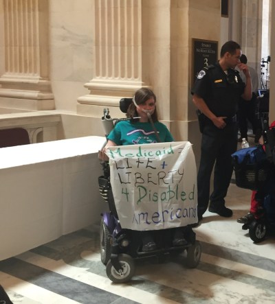 Laura Halvorson in her bipap breathing mask and power wheelchair wearing a turquoise ADAPT Disability Integration Act shirt. Next to her is another ADAPTer and police officers getting ready to exit the Dirksen Senate Building after getting arrested on June 22, 2017 outside Mitch McConnell’s office. She is holding a banner that says Medicaid = Life + Liberty 4 Disabled Americans.