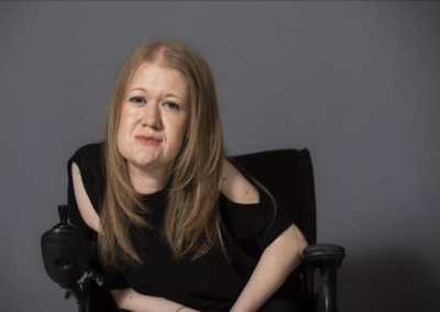 Picture is of Frances sitting in her wheelchair; smiling, wearing a black top and her blonde hair down.