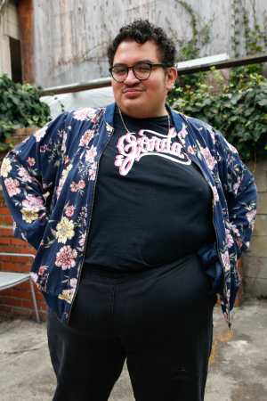 Caleb, a fat brown disabled femme, places their hands on their hips and looks at the camera defiantly. They are wearing blue plants, a blue bomber jacket with pink florals, and a black t-shirt that reads GORDA in a cursive script.