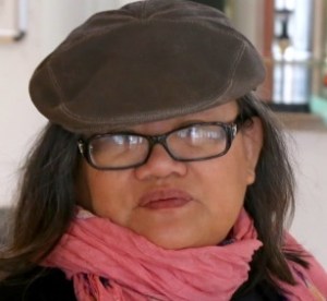 Middle-aged Asian woman with shoulder-length long hair, a hat on her head, and eyeglasses. She has a pink scarf wrapped around her neck.