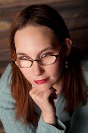 Elsa Sjunneson-Henry, a caucasian woman with long red hair, wears glasses which she is looking over. One eye is hazel, the other is a white occuluded cataract. She wears a teal sweater, pink collared shirt, and a smile that says "you want me to write words for you, right?"