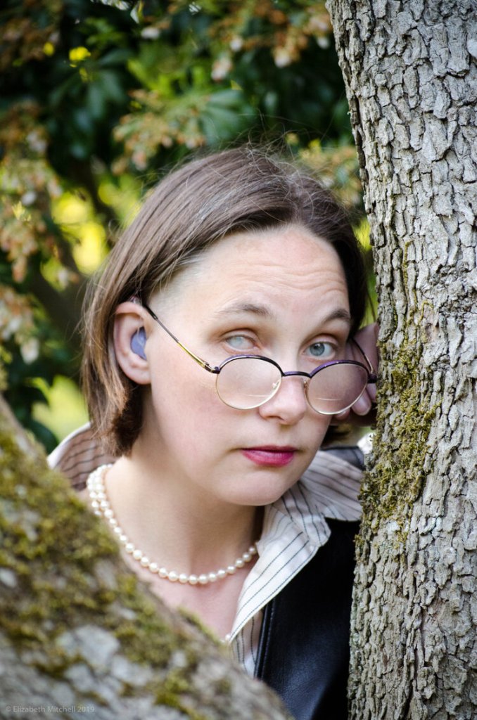 A white woman with short hair and an occluded cataract on her right eye wearing purple hearing aids and a pearl necklace, looking with raised eyebrows at camera between trees. Photo credit: Lis Mitchell, 2021.