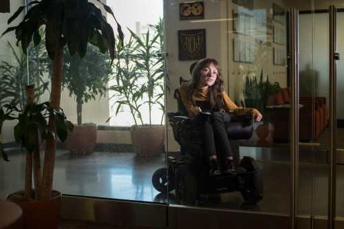 Emily sits among plants in a power wheelchair behind a glass door. She is dimly lit, with long brown hair, a tan turtleneck, and black pants and boots.