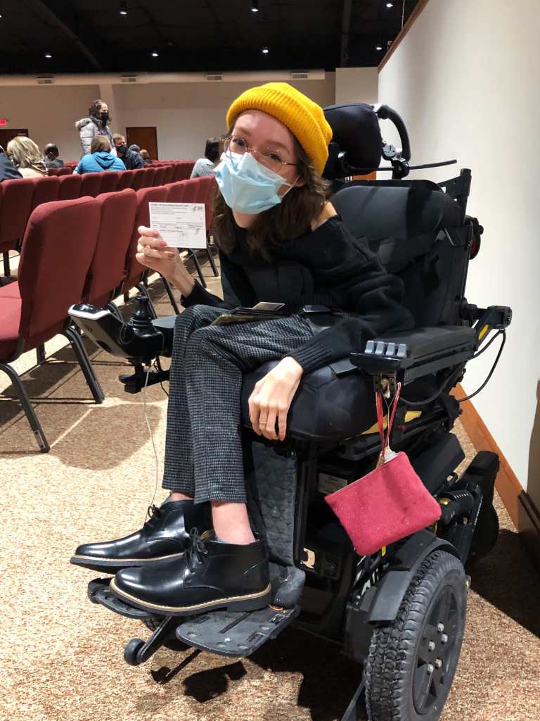 Emily Ackerman, a white woman in a black power wheelchair is holding up her COVID vaccine card. She’s wearing a black sweater and boots with grey checked pants, a yellow beanie, and a blue medical mask.