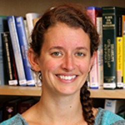 Young white woman with long brown hair tied in a braid. She is smiling at the camera and standing in front of a bookshelf full of books.