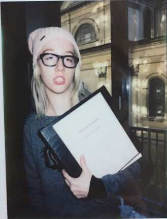 A young Caucasian woman with short blonde hair wearing a light pink beanie, a grey sweatshirt, and dark green glasses, standing in front of a window and holding a large binder.