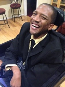 Photo description: Lateef McLeod is smiling and winking, at the camera in his black suit. He is sitting in his power wheelchair and his tray and headrest are visible. To the side of them stools, a hardware floors, and an open doorway. Behind him is a tan bookshelf and a maroon couch.