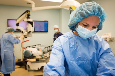 Photo of a surgical bay. In the foreground is a woman in a blue gown, face mask and cap over her head. In the background are other doctors and someone laying in a hospital table. 